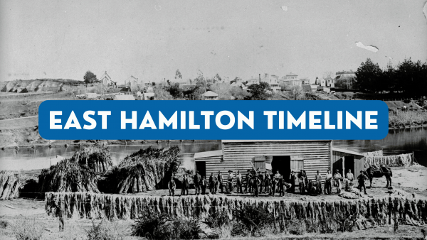Black and white image showing staff of a flax mill gathered in front of the building. The building sits on a riverbank. A blue and white title is overlaid over the image reading East Hamilton Timeline.