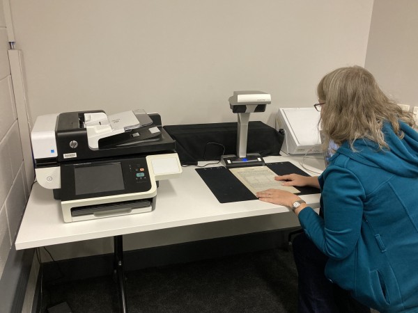 A colour image showing a flatbed scanner and overhead scanner sitting on a white desk. A woman in a blue jacket sits at the desk placing a sheet of paper under the overhead scanner.