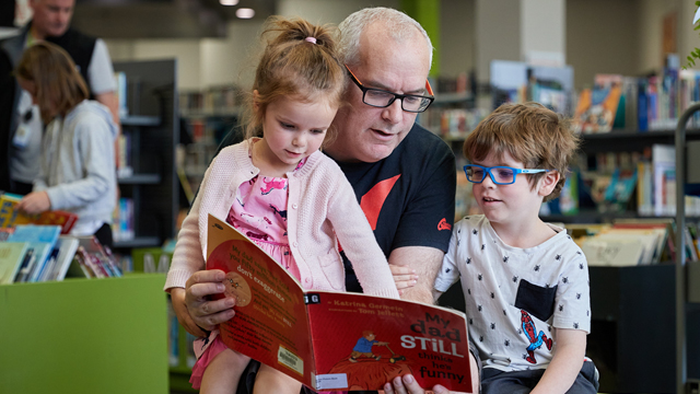 Image of a father reading a picture book to his son and daughter at Central Library in the children's area.