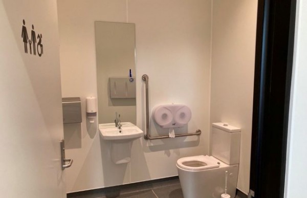 The accessible toilet/family room door open, showing a wall mounted hand dryer, hand basin, handrail on the wall next to the toilet. In the mirror above the hand basin is the reflection of a baby changing table mounted on the opposite wall.