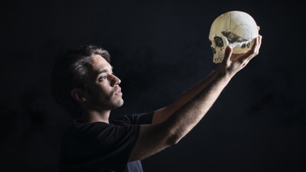 A man standing in the dark, holding a skull high above and staring at it sadly.